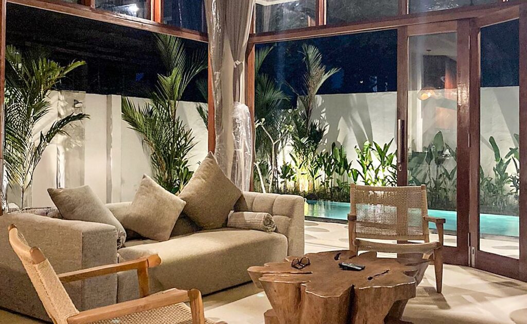 The living room of Villa Ceria at night: sofa, rattan armchairs and a teak root table, with the folding doors open to the lit pool and palms