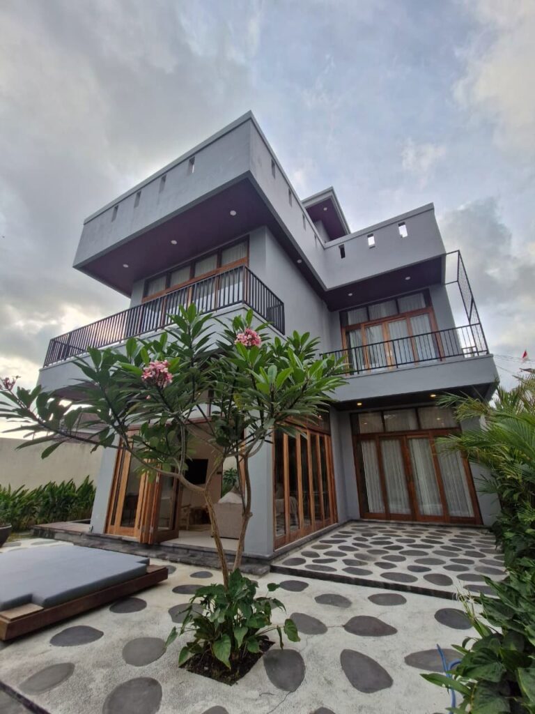 Villa Ceria from the garden: the house with balconies and teak folding doors, a frangipani tree and stepping stones in front